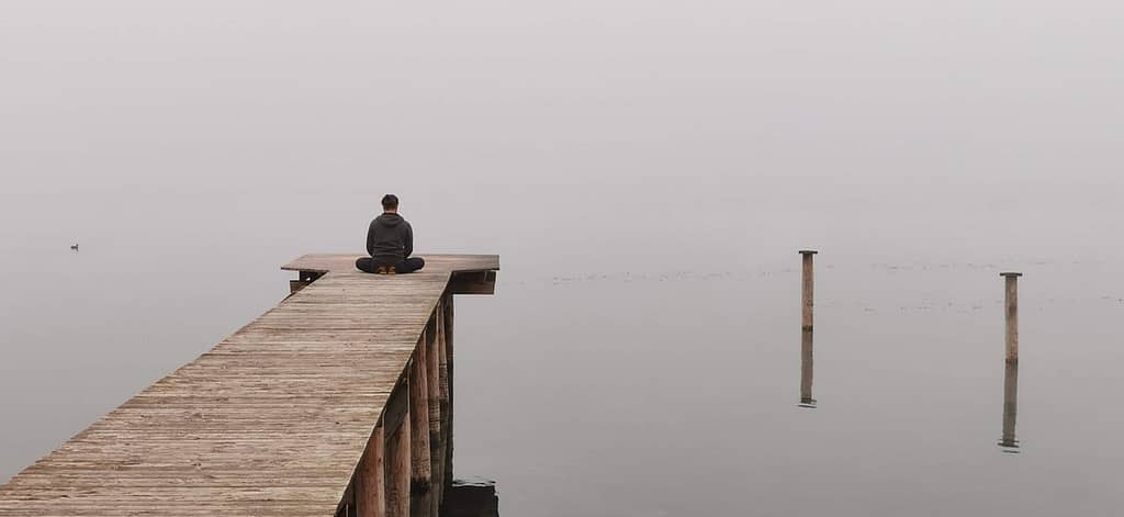 Man sitting in lotus position at the end of a wooden pier over the ocean, using his shoes as a cushion, facing the foggy and cold water, symbolizing peaceful reflection and meditative states.