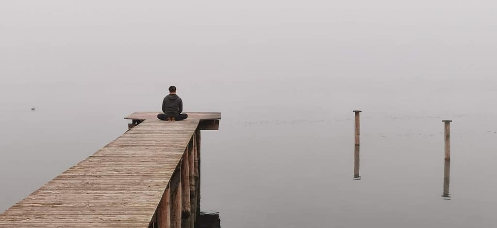 Man sitting in lotus position at the end of a wooden pier over the ocean, using his shoes as a cushion, facing the foggy and cold water, symbolizing peaceful reflection and meditative states.