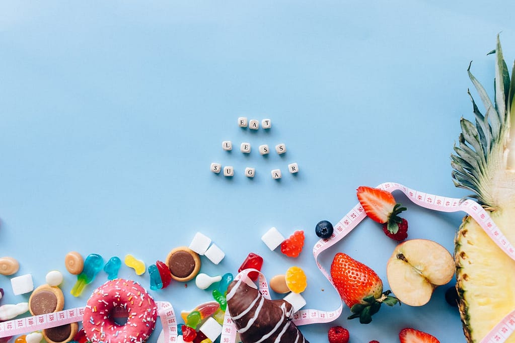 On a light blue background, the words 'eat less sugar' are spelled with letters resembling dice. Below, sugary foods like cookies, doughnuts, gummy candies, fruits, and sugar cubes are crowded together with a tape measure wrapped around them.