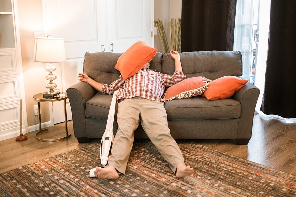 Man lying half on the floor, half on a sofa, with a pillow over his head and a vacuum cleaner by his side. His legs are stiff and straight, suggesting he fell asleep mid-cleaning.