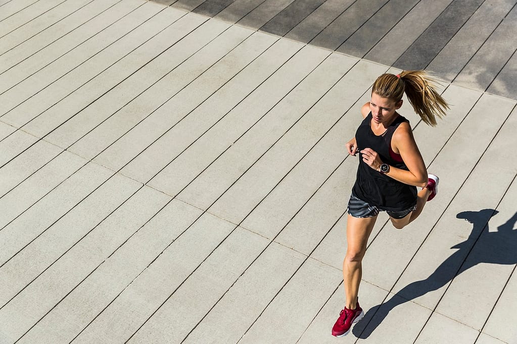 Fit woman running on pavement designed to look like wooden boards, shot from an above-side angle. She wears typical running gear with her hair in a ponytail, focused on her stride as she runs on a clean concrete surface resembling a wooden deck.