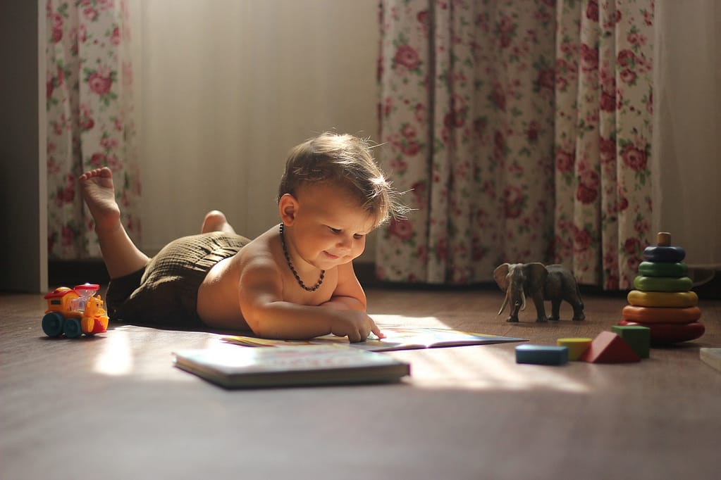 Smiling baby, around 1 year old, lying on tummy, looking at a book with a toy train, dinosaur, shapes, and blocks surrounding them, engaging in developmental play.
