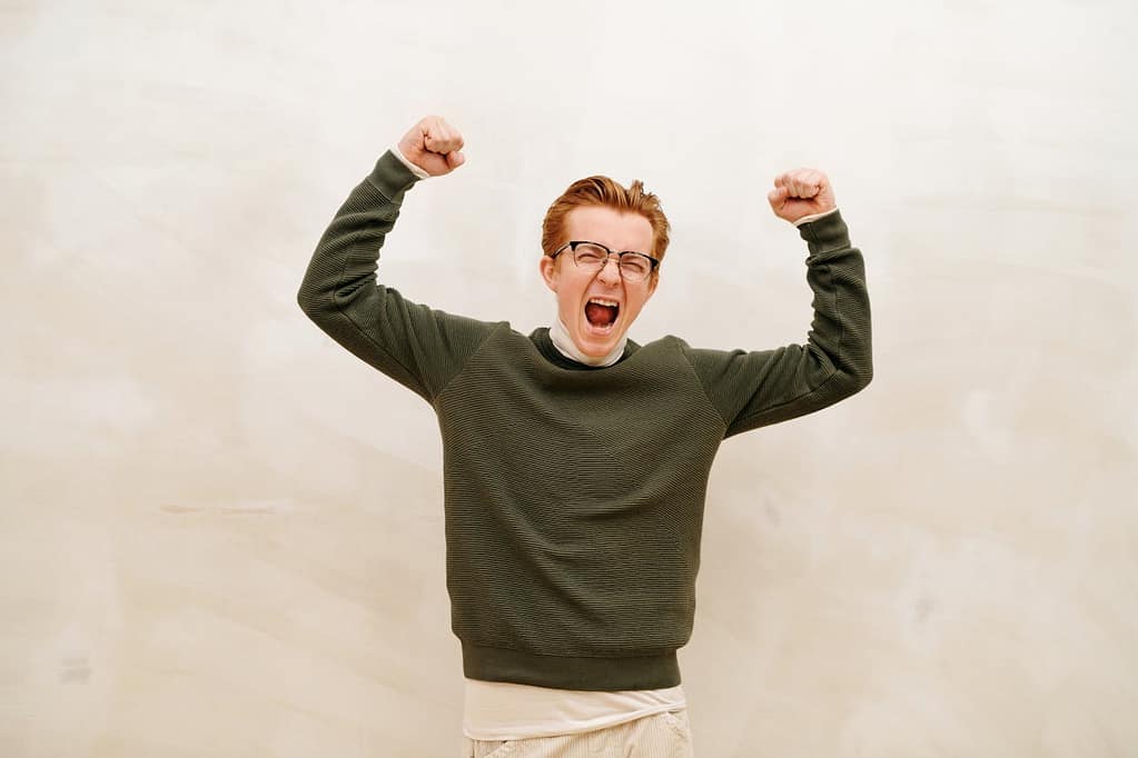 A young man wearing glasses and a green wool sweater, cheering with both arms raised in excitement, set against a clean white/beige background.