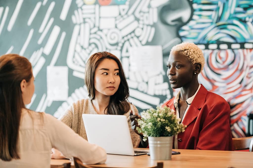 Three women working together around a table, with two of them providing feedback to the third women.