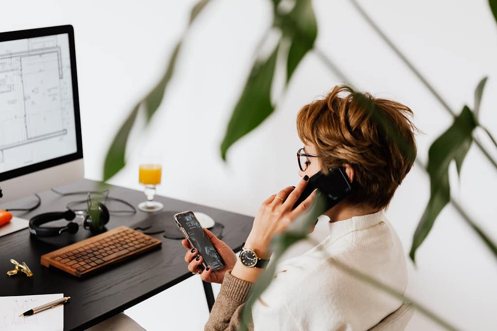A woman multitasking at her desk, sitting in front of her computer with a design open on the screen, talking on one phone while scrolling on another, with headphones and a glass of orange juice on the table.