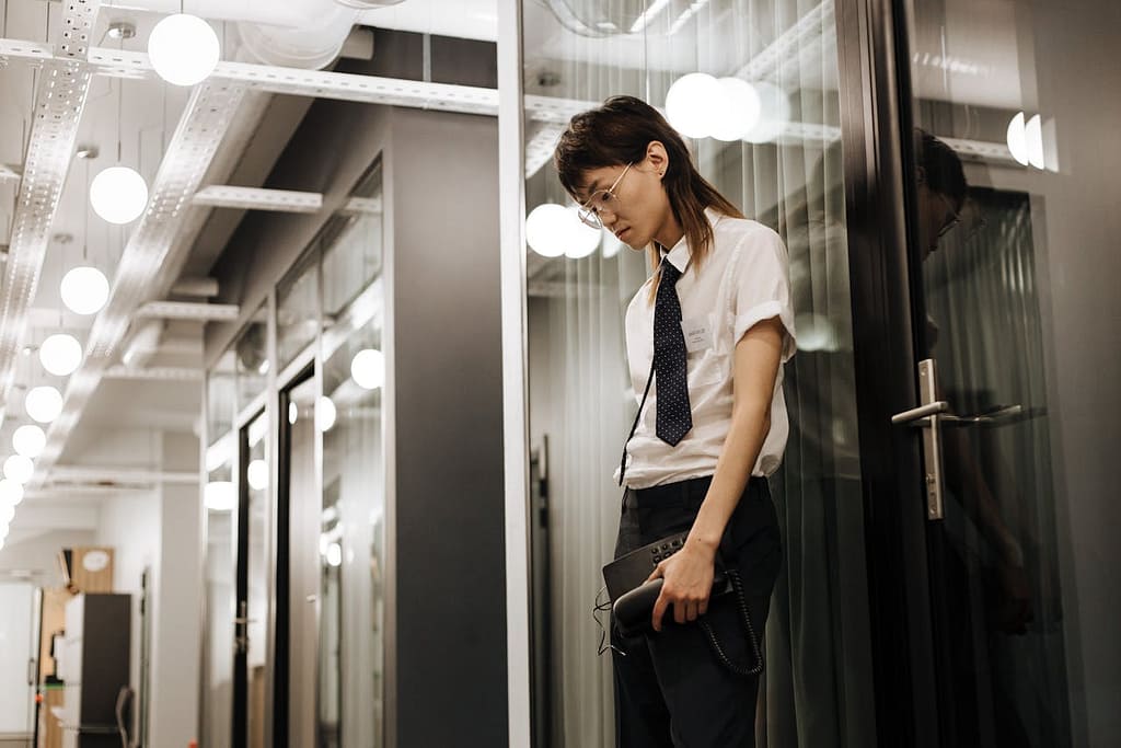 Young Asian man in smart business attire, holding a phone in a corporate building, looking down with a stressed expression. His mullet hairstyle contrasts with the formal setting.