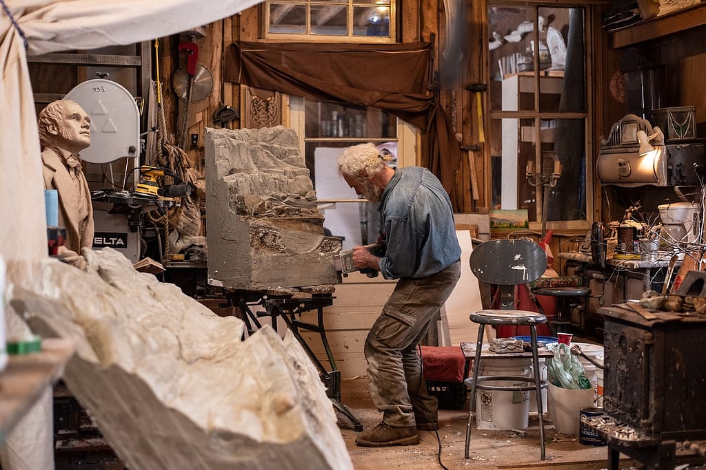 An elderly man working on a sculpture in his art studio, surrounded by tools, paint cans, and half-finished projects.
