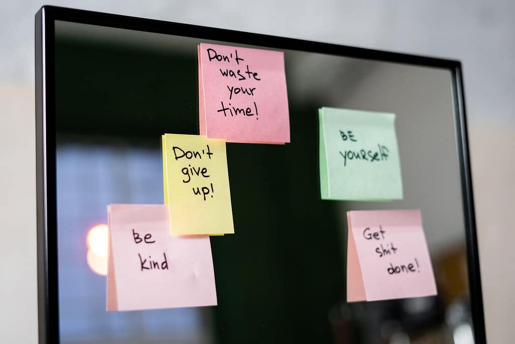A glass mirror covered with colorful sticky notes containing motivational messages like "Be yourself," "Get shit done," and "Don't give up."