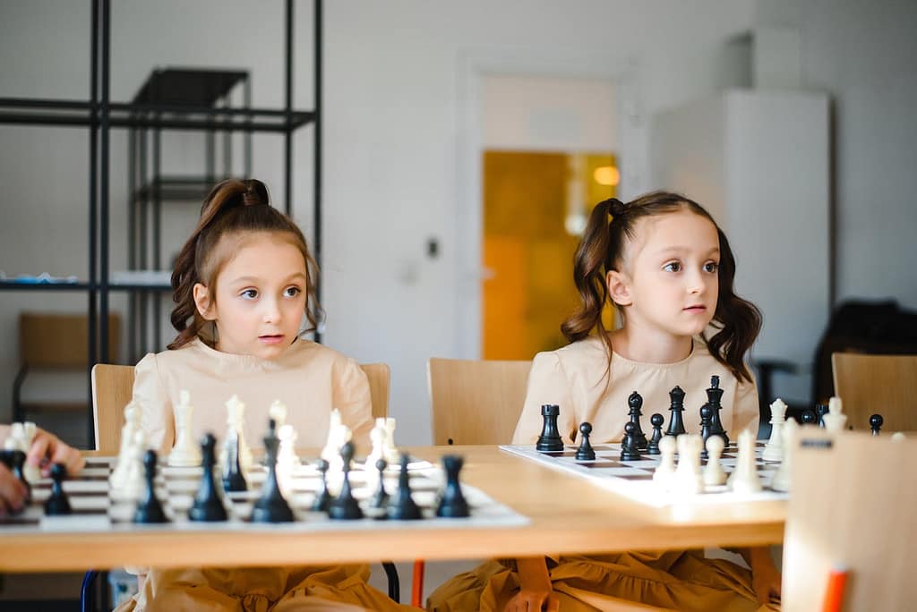 Identical twin girls with matching beige dresses sitting at separate chess boards.