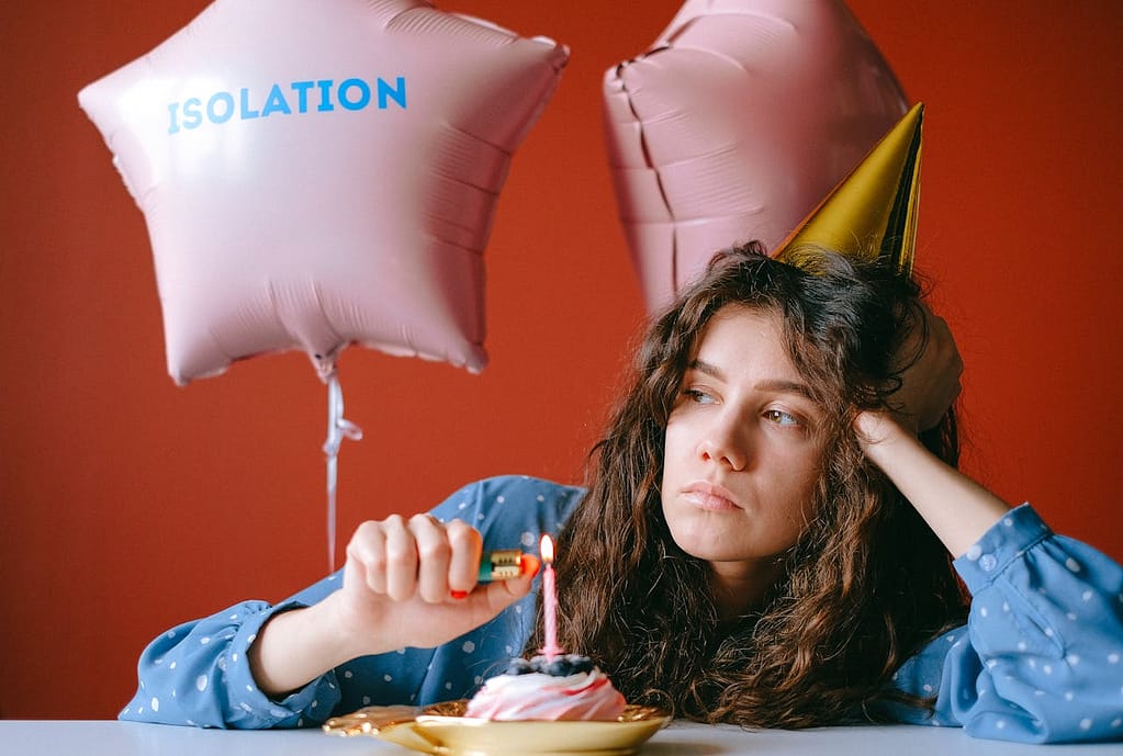 Woman sitting alone at her birthday with a cupcake and single candle, wearing a birthday hat, looking bored with balloons behind her, one reading 'isolation.