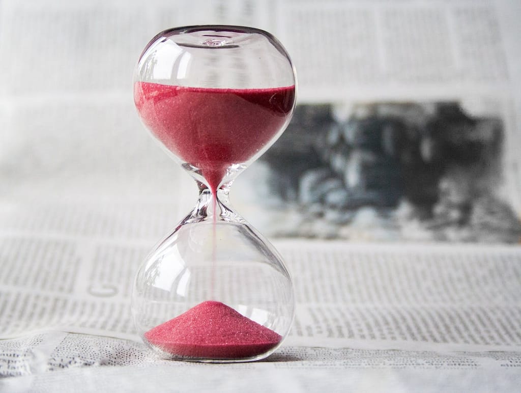 An hourglass with red sand, one-quarter filled, standing on a plain newspaper background.