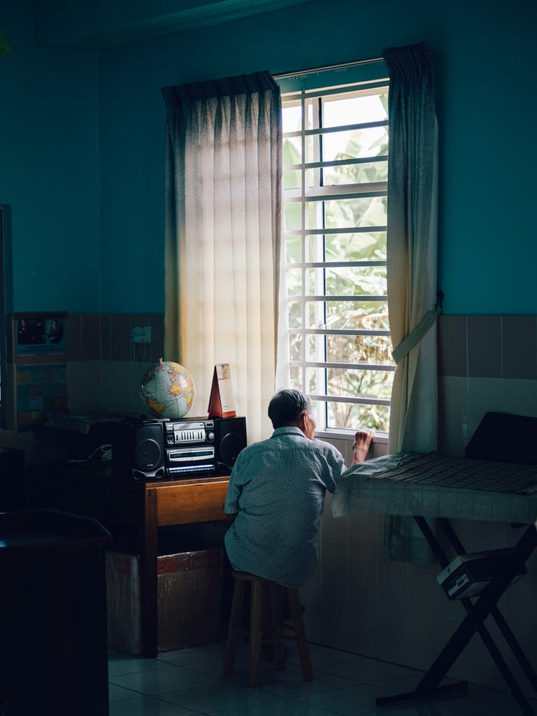 Older man sitting on a stool in his kitchen, gazing out the window during lockdown, representing isolation and disconnection.