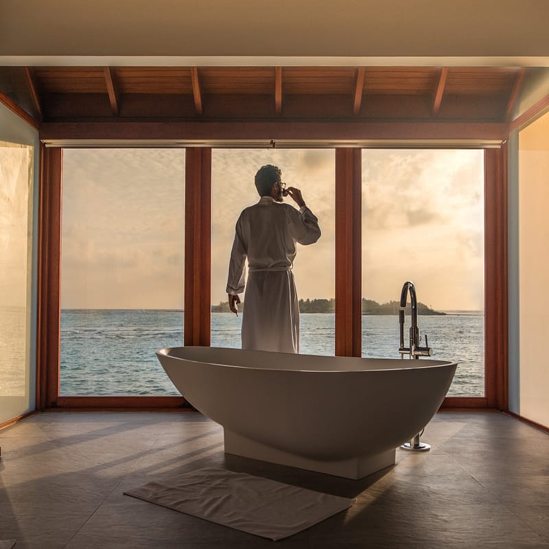 Man in a robe sipping coffee while standing in a luxurious bathroom with a glass wall, overlooking the ocean.